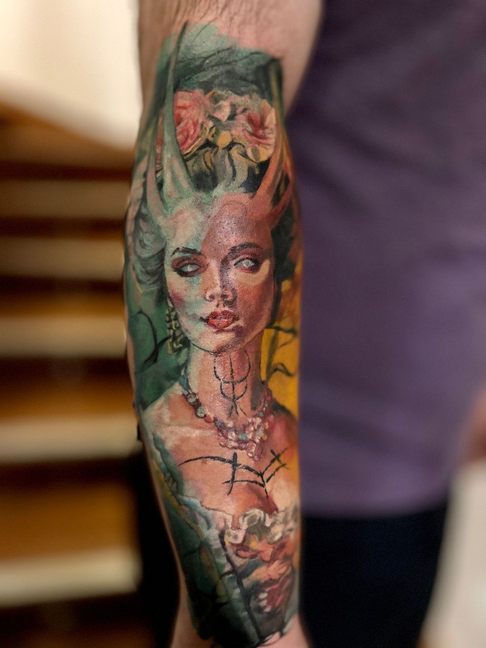 Tattoo of a woman's portrait, with horns and ornate jewelry, in watercolor style on an arm.