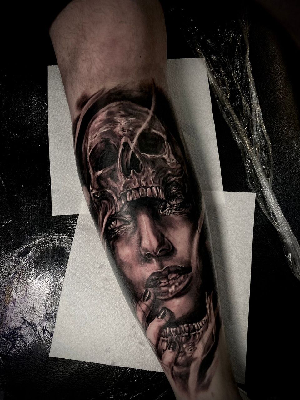 Tattoo of a skull merging with a woman's face on an arm, in black and gray ink.