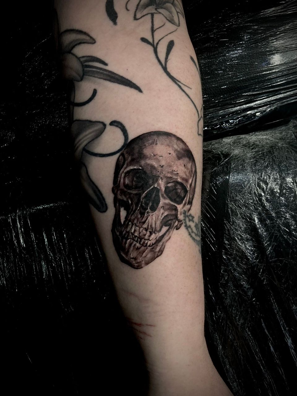 Tattoo of a skull on an arm, with floral designs partially visible. Dark, detailed shading.