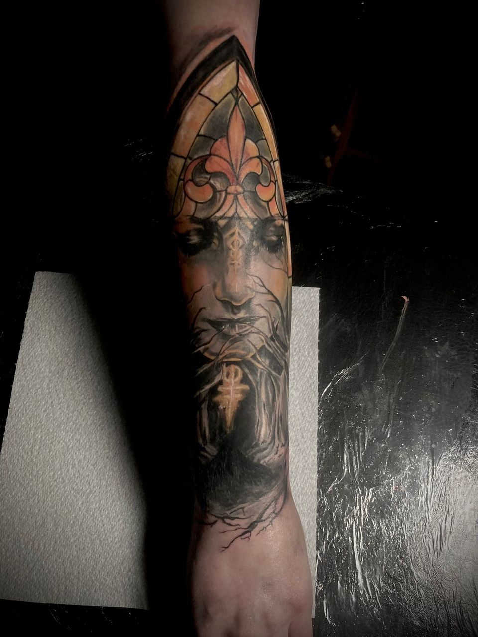 Detailed tattoo on an arm, featuring a face with a fleur-de-lis and stained glass window design in yellow and black.