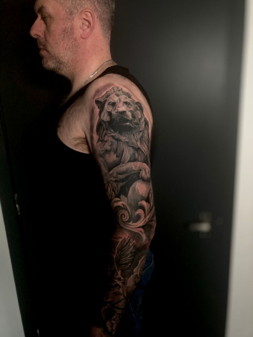 Man with full sleeve tattoo featuring a lion statue design, in a dark room.