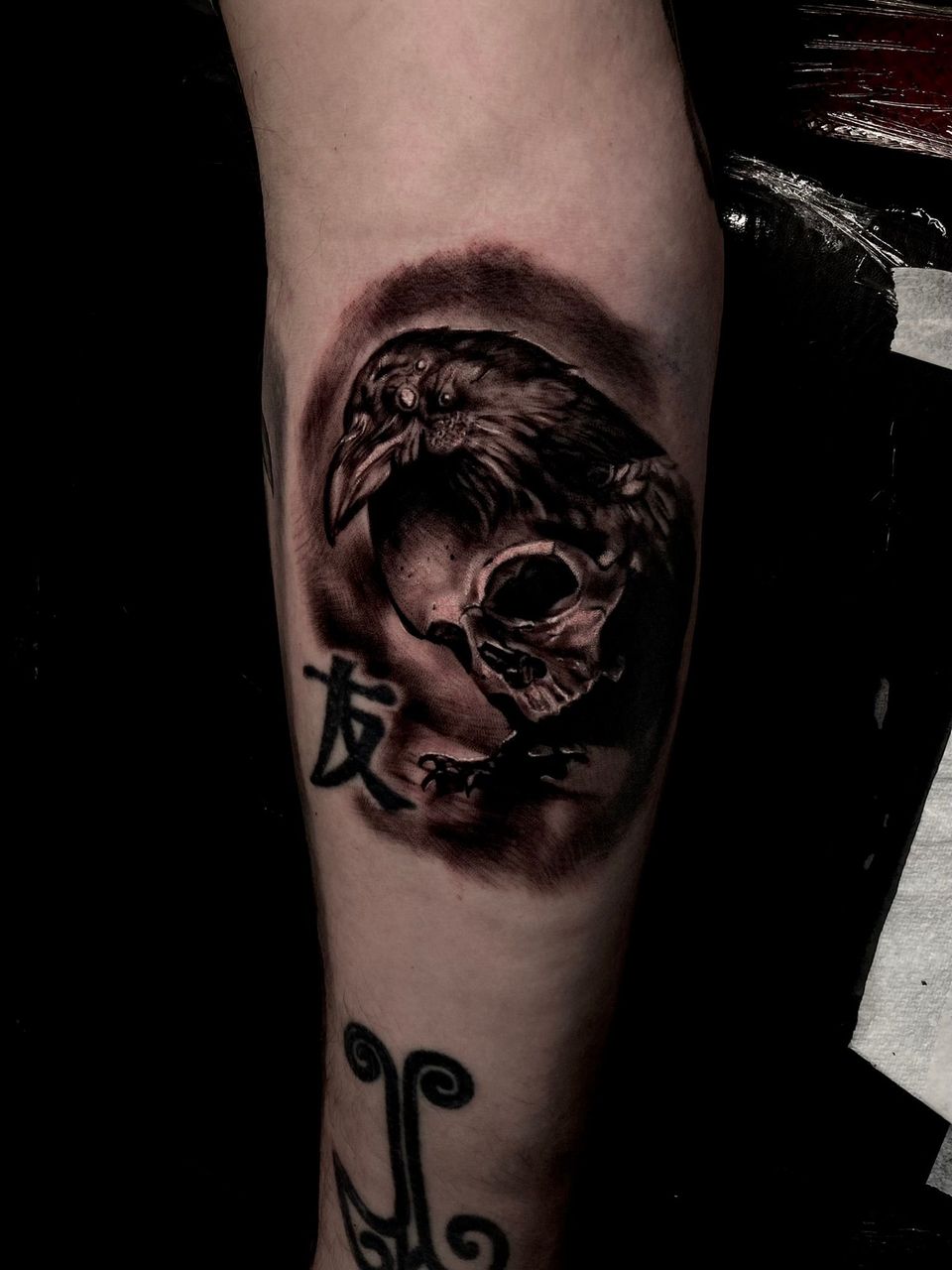 Tattoo of a raven perched on a skull; surrounded by dark shading. Chinese symbol 