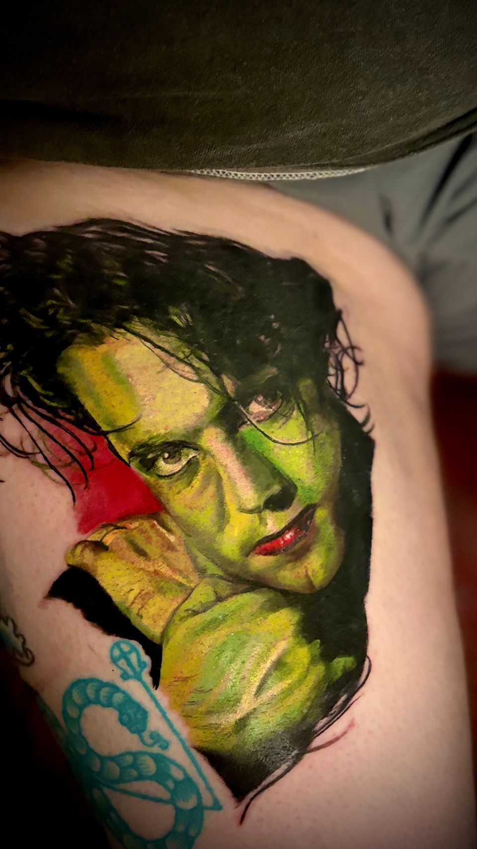 Tattoo of a person with green skin, black hair, and red lips, in a portrait with a red background.