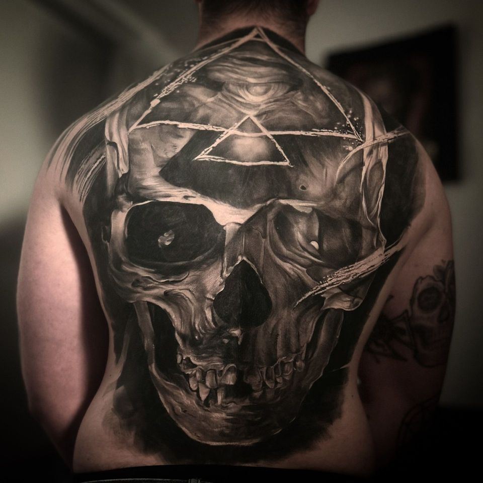 Back tattoo of a detailed skull with geometric shapes. Black and gray shading with visible teeth and eye sockets.