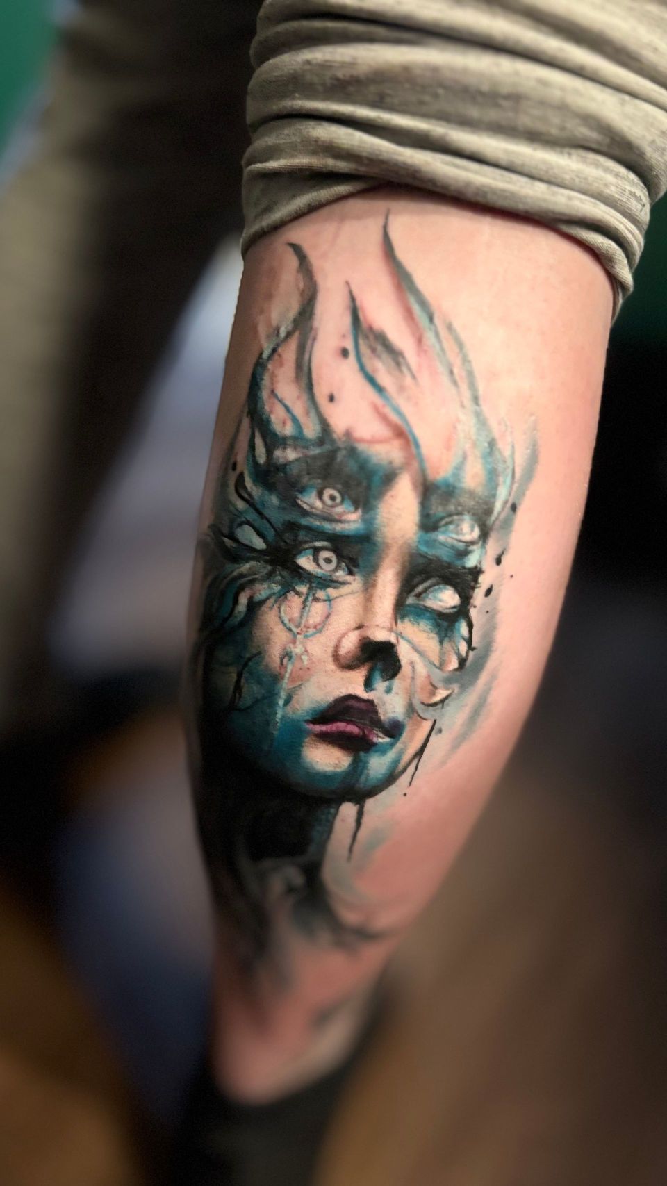 A tattoo of a pale face with a smoky, blue-green aura. The eyes are white. The lips are deep purple.