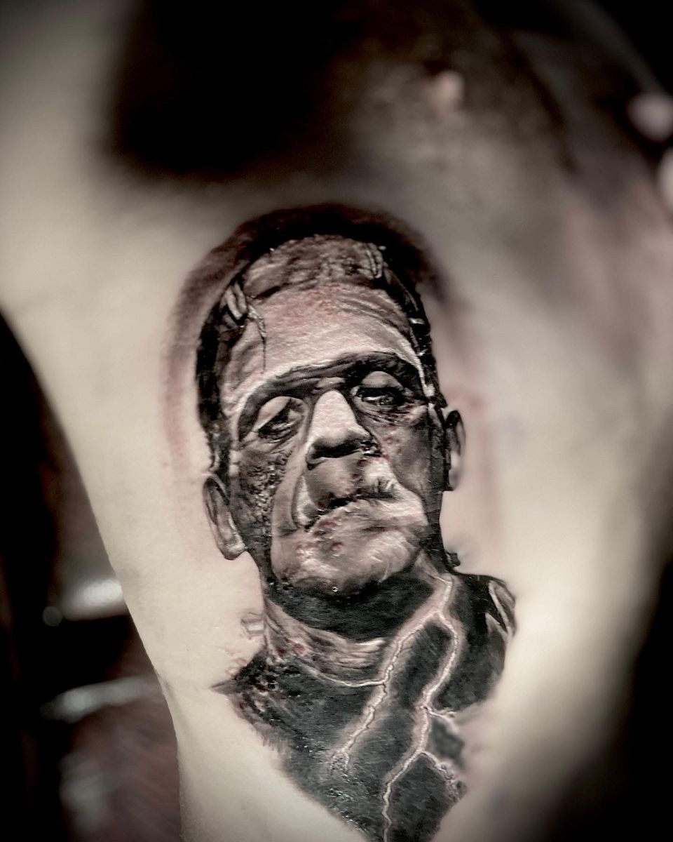 Frankenstein's monster tattoo on a person's side in black and gray. Features stitches, scars, and a dark shaded background.
